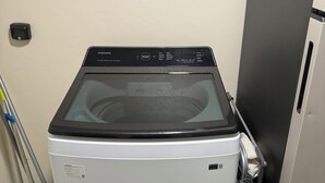 Une machine à laver compacte, de couleur argentée, dotée d’un panneau de commande numérique, placée dans un coin d’une pièce dont le sol est carrelé et où un balai est appuyé contre le mur.