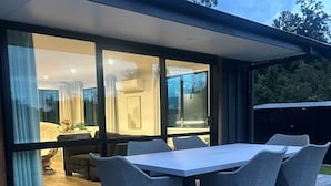 A modern outdoor dining area with a table and chairs, a wooden deck, and a view of the interior through glass doors.