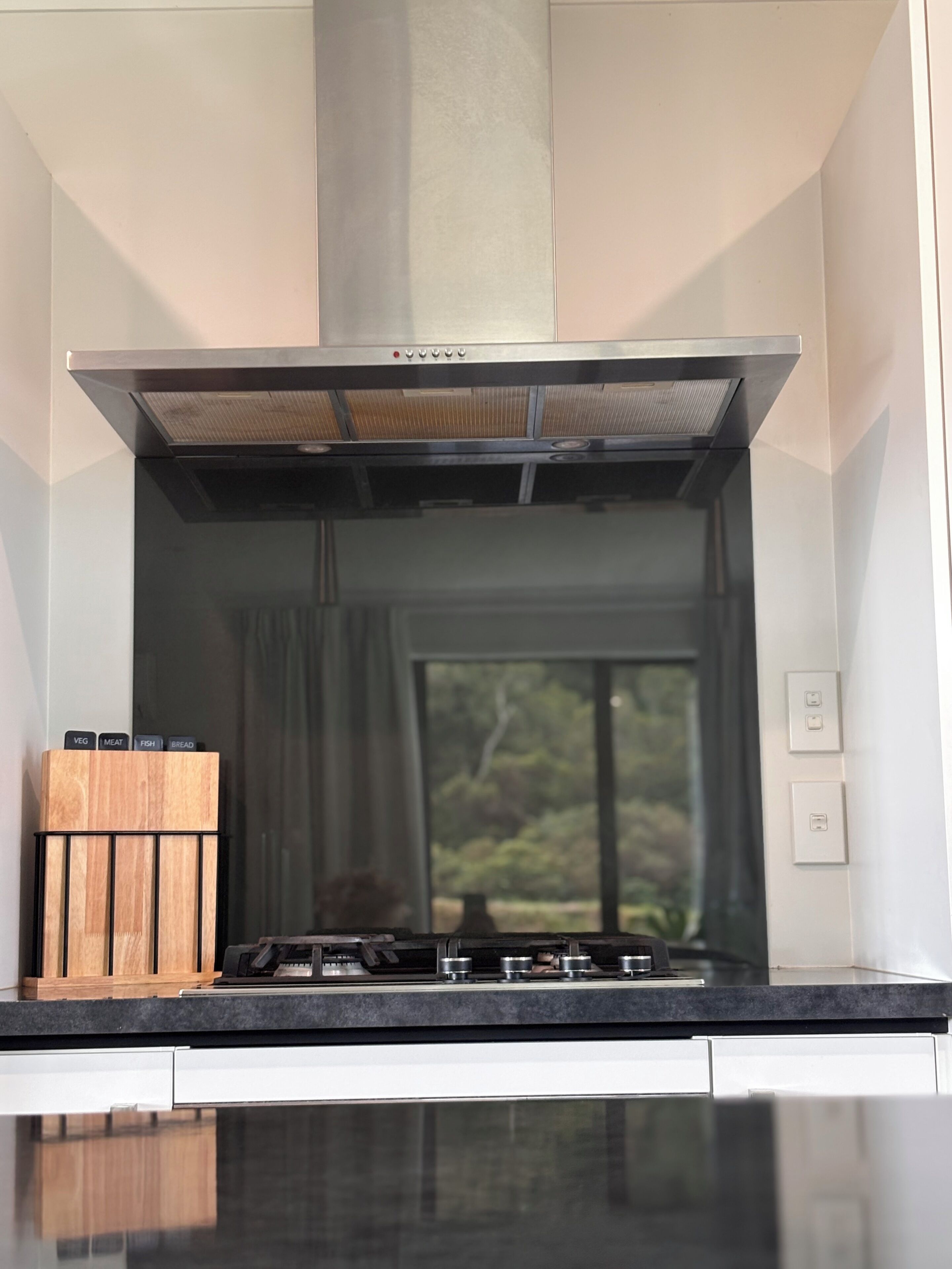 A modern kitchen with a stainless steel range hood, a wooden cutting board, and a window with a view of greenery.