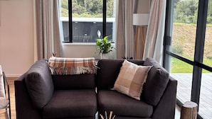 A modern living room with a dark-colored sectional sofa, a coffee table with a candle, and a view of greenery outside.