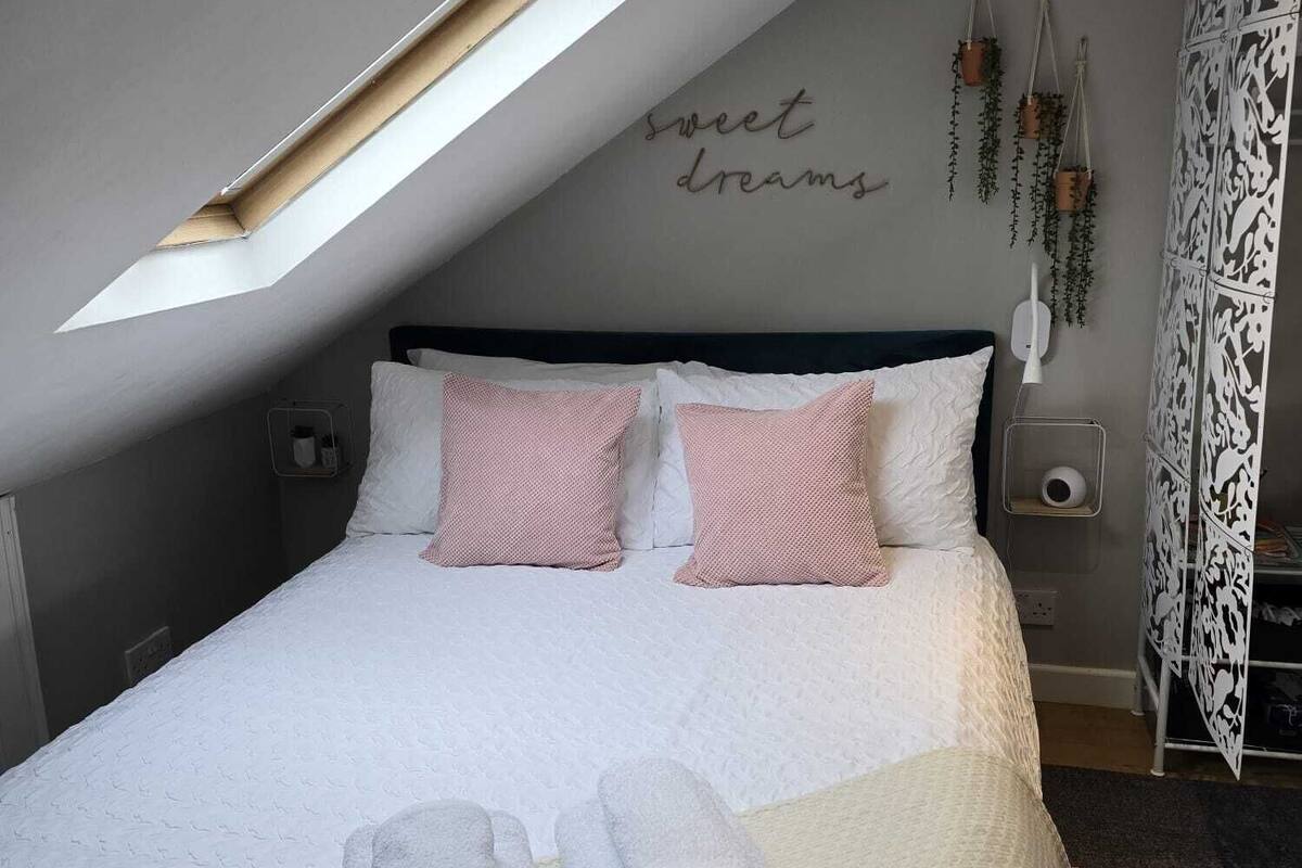 A bedroom with a skylight, a bed with pink and white pillows, a bedside table with a lamp, and a wall with a 'sweet dreams' sign.