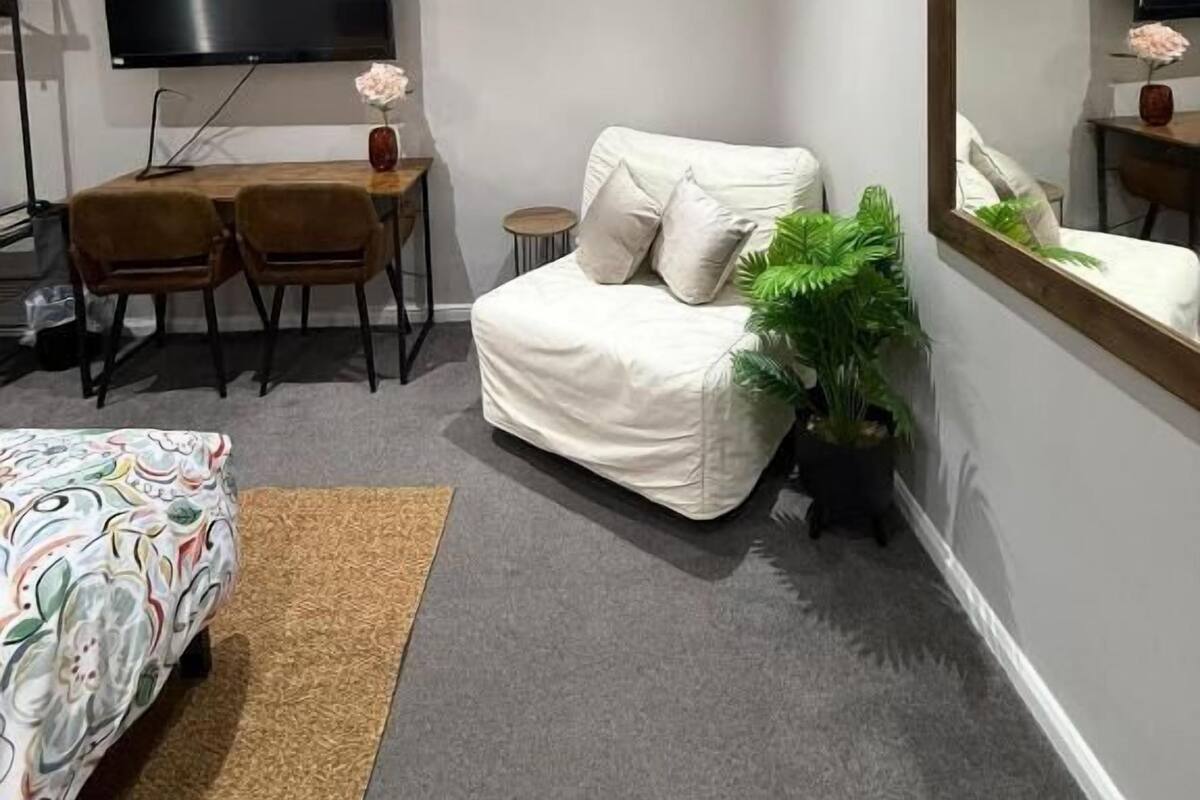 A modern hotel room with a bed, a sofa, a dining table with chairs, a flat-screen TV, a mirror, and a potted plant.