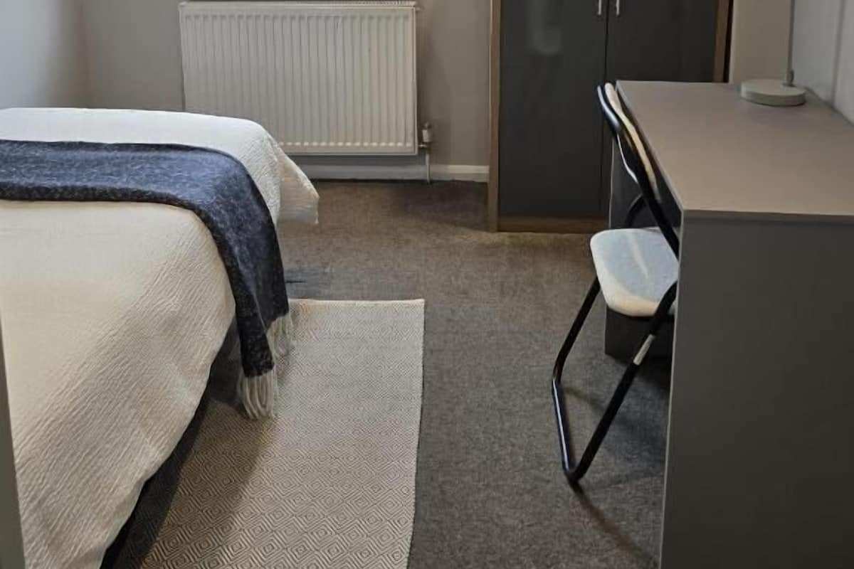 A bedroom with a bed, a desk, a chair, a wardrobe, and a window with blinds.