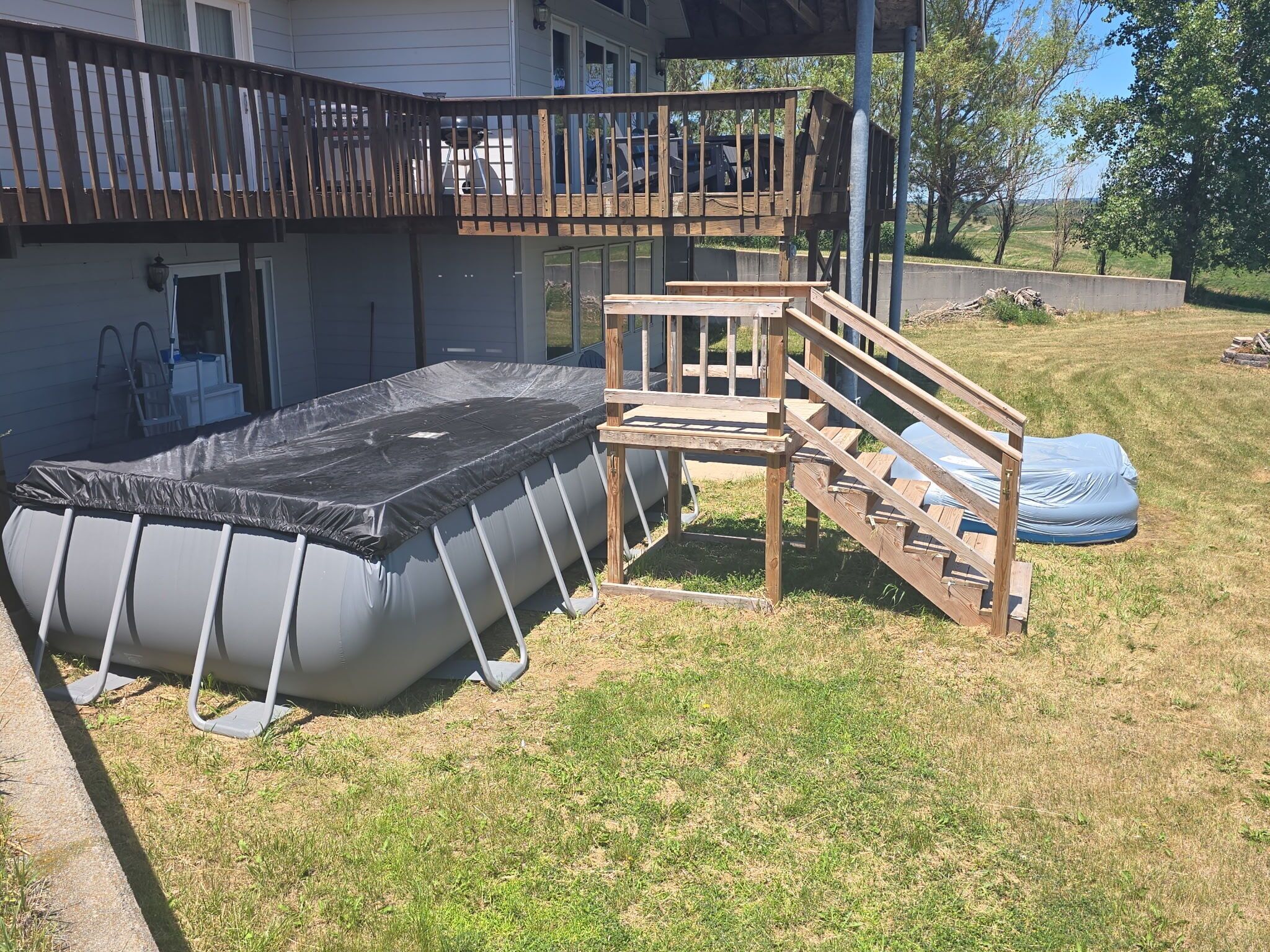 Een opblaasbaar zwembad met een houten terras en trap, een overdekte patio en een huis met balkon.