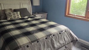 Un dormitorio con cama, mesita de noche, lámpara, cuadro enmarcado y ventana con vista a árboles.