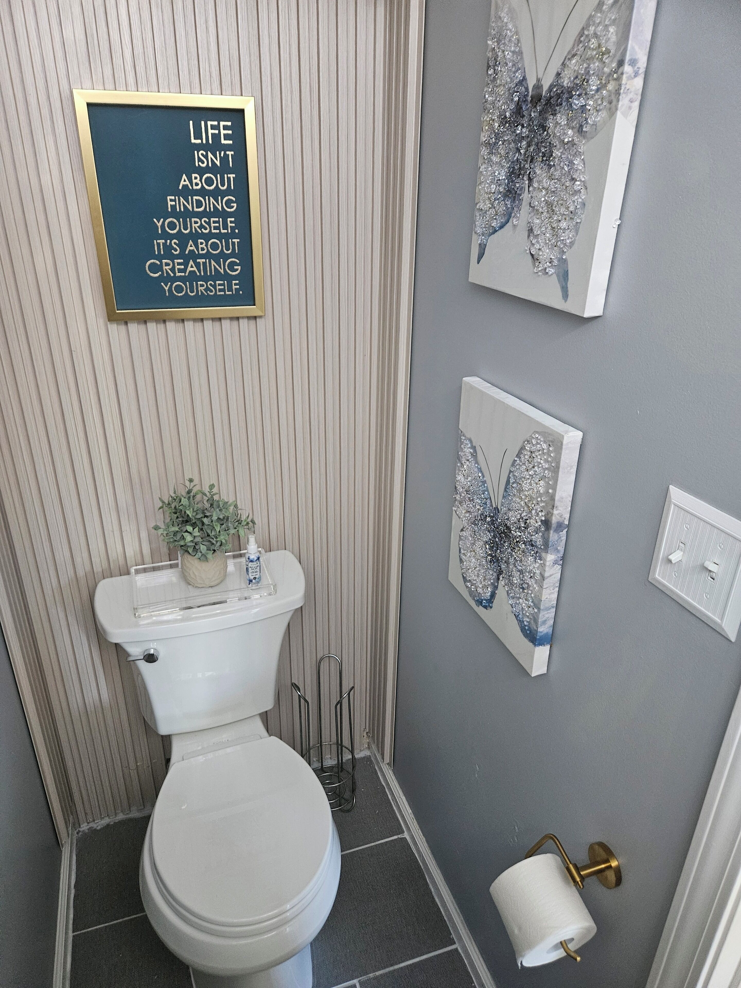 Une salle de bain avec des toilettes, une citation inspirante encadrée et deux œuvres d’art représentant des papillons accrochées au mur.