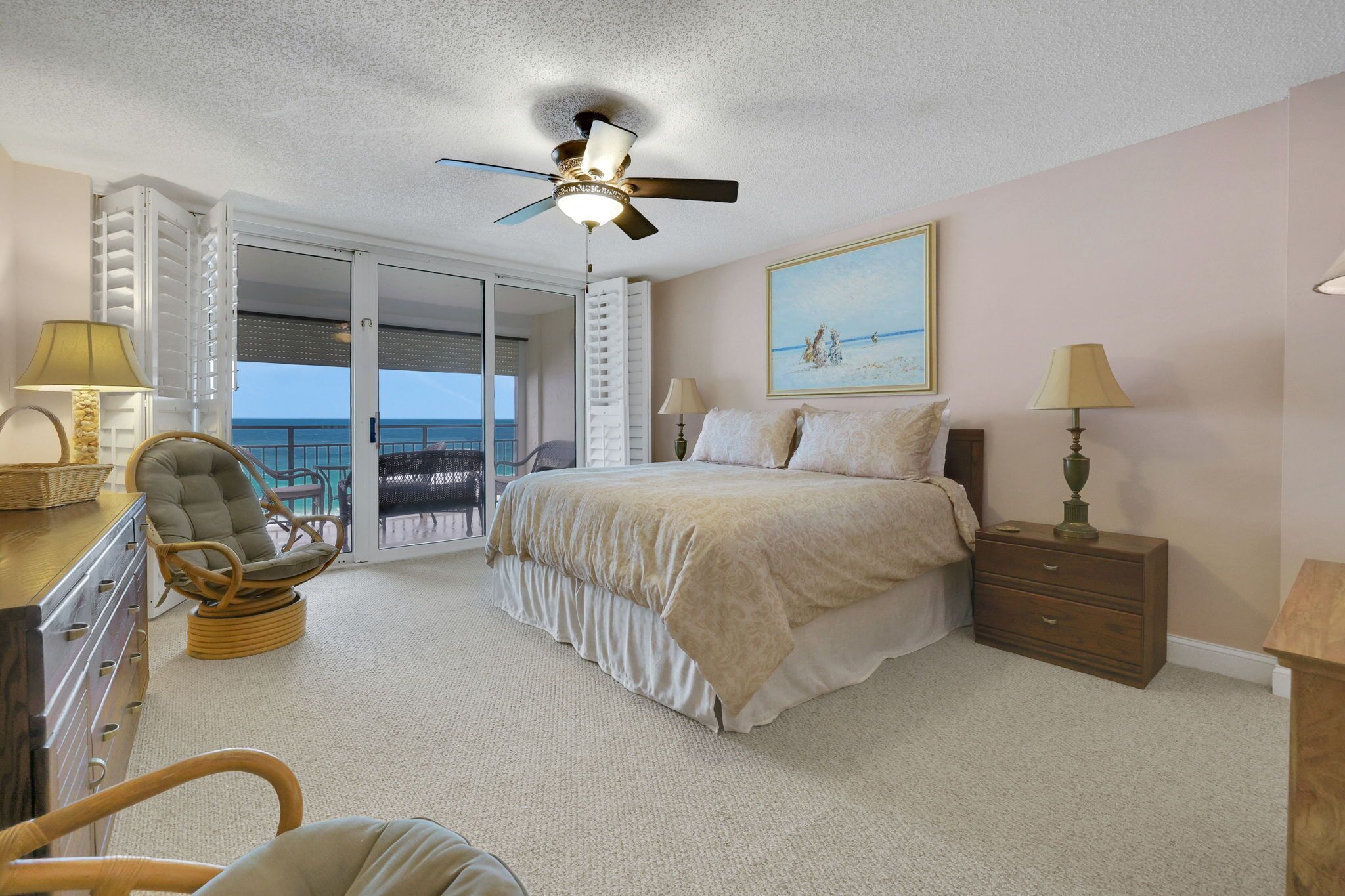 A bedroom with a bed, a ceiling fan, a nightstand, a lamp, a chair, and a view of the ocean through a sliding door.