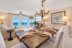 A hotel room with a wooden dining table set for two, a sofa, a chair, a ceiling fan, a chandelier, a TV, and a view of the ocean. - Oceanfront Luxury Condo | Renovated | Private Beach Access | Pools | Tennis Court (St. Augustine)