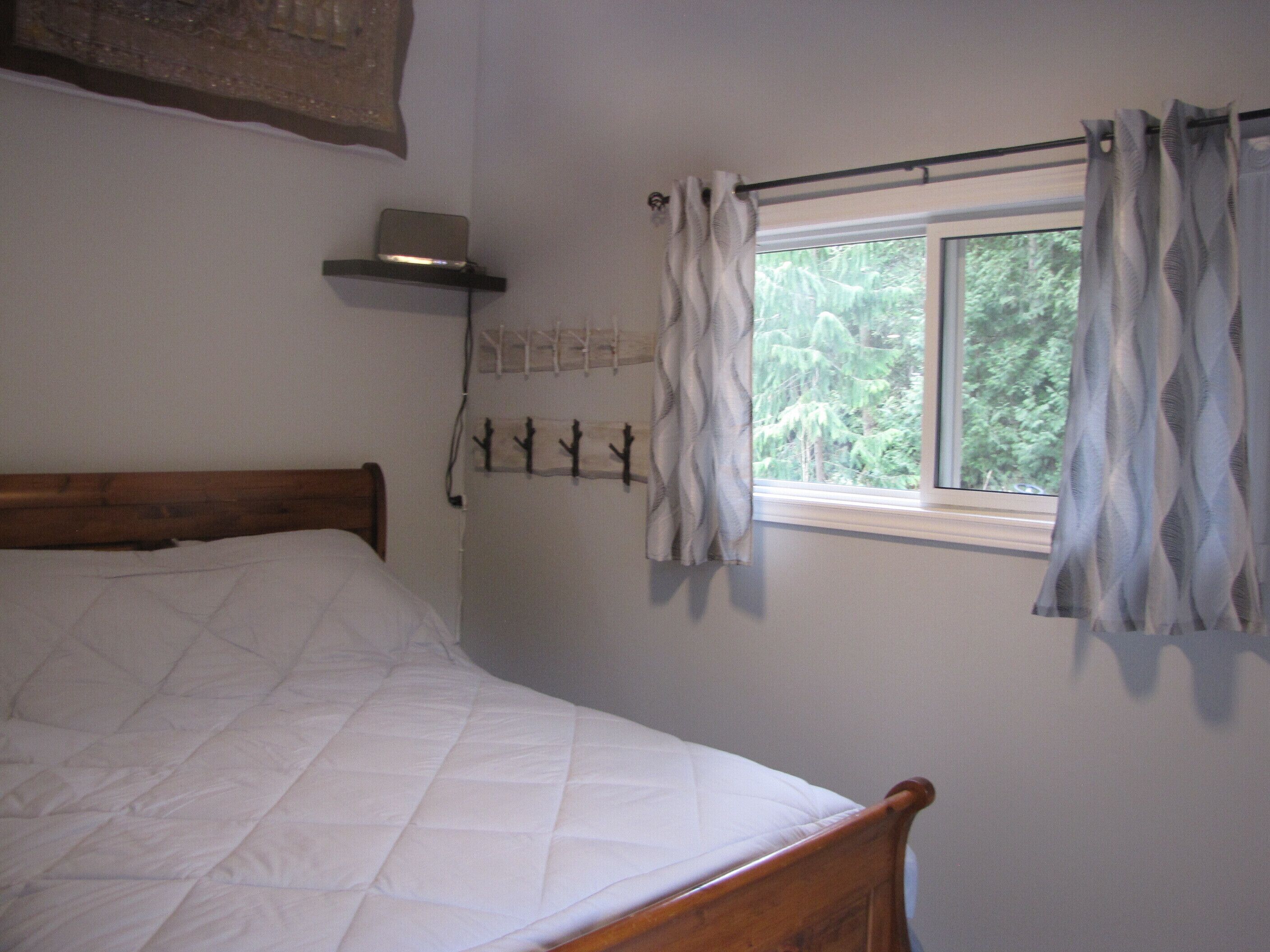 A single bed with a wooden headboard, a window with curtains, a wall-mounted shelf with a laptop, and a coat rack with hooks.