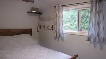 A single bed with a wooden headboard, a window with curtains, a wall-mounted shelf with a laptop, and a coat rack with hooks.