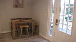 Una habitación con una mesa de madera, dos taburetes, una puerta con ventanal y una obra de arte colgada en la pared.