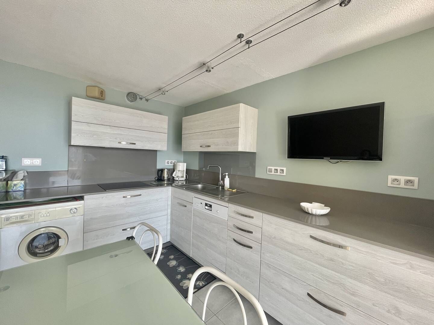 A modern kitchen with a built-in washing machine, a dining table, and a flat-screen TV mounted on the wall.