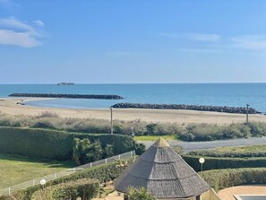 Vista da praia com uma construção de colmo, um caminho pavimentado e um navio ao longe.