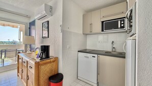 A compact kitchen with a microwave, dishwasher, and a small sink. There is a wooden cabinet with a red trash bin, and a window offering a view outside. - CAP D'AGDE - Plage Richelieu / Réf 307 (CAP D'AGDE)