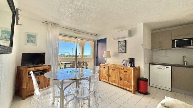 A modern kitchen with a microwave, sink, and dishwasher, a dining area with a glass table and chairs, and a view through a glass door. - CAP D'AGDE - Plage Richelieu / Réf 307 (CAP D'AGDE)