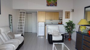 Ein Wohnzimmer mit einem weißen Sofa, einem Glastisch, einem weißen Sessel, einer Leiter, einem Küchenbereich mit Schränken, einem Kühlschrank und einem Gemälde an der Wand.