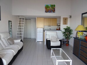 Ein Wohnzimmer mit einem weißen Sofa, einem Glastisch, einem weißen Sessel, einer Leiter, einem Küchenbereich mit Schränken, einem Kühlschrank und einem Gemälde an der Wand.