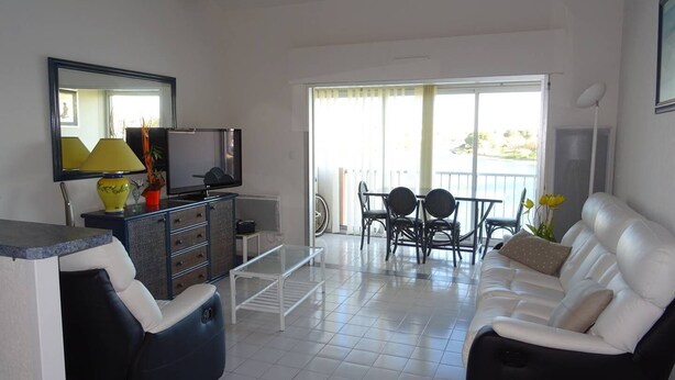 Ein Wohnzimmer mit einer Couch, einem Fernseher, einem Esstisch mit Stühlen, einem Balkon mit Blick und einer Decke mit sichtbaren Balken.