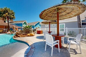 A pool area with a table and chairs, a thatched umbrella, and a white fence. - PB117 Shared Pool, Pet Friendly, Parking for 3, Charcoal Grill (Port Aransas)
