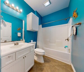 A bathroom with a white sink, mirror, and vanity, a toilet, and a bathtub with a shower curtain. - PB117 Shared Pool, Pet Friendly, Parking for 3, Charcoal Grill (Port Aransas)