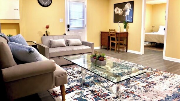 Ein Wohnzimmer mit Sofa, einem Glastisch, einem Teppich mit Muster, einem Essbereich mit Tisch und Stühlen sowie einem Schlafzimmer, das durch eine geöffnete Tür sichtbar ist.