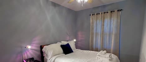 Une chambre à coucher avec un lit, une table de chevet, un ventilateur de plafond, une fenêtre avec des rideaux et un plancher en bois.