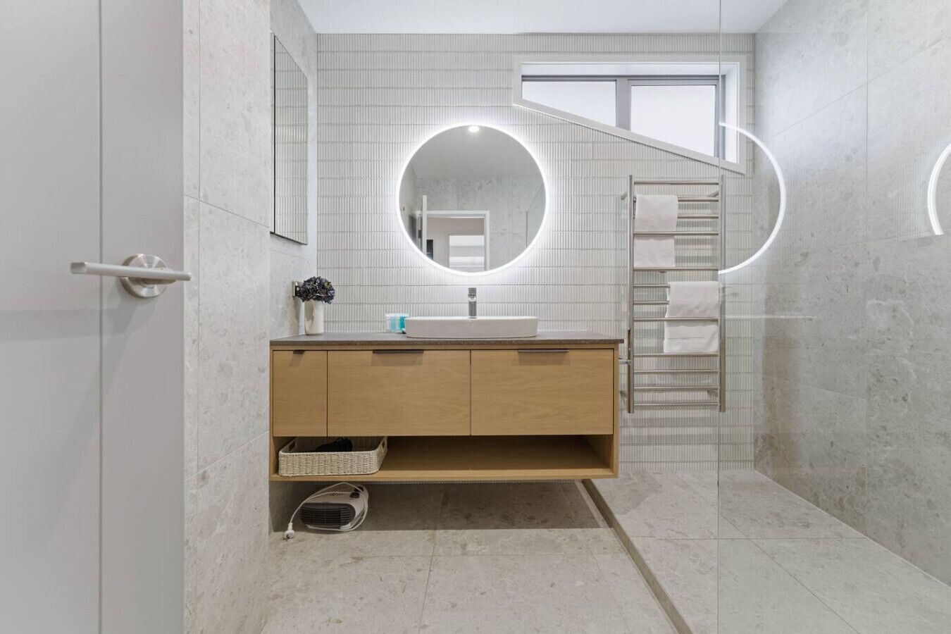 A modern bathroom with a wooden vanity, a mirror with a circular light, a towel rack, and a shower area.