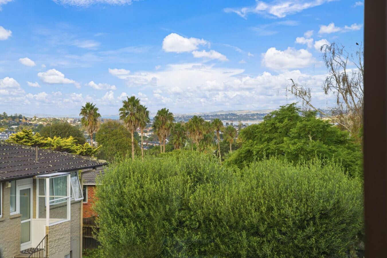 A residential area with a house, dense greenery, and a distant cityscape under a partly cloudy sky.