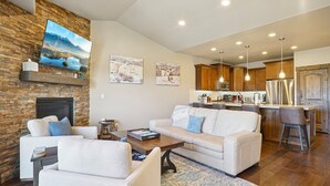 A modern living room with a fireplace, a sofa, a coffee table, and a kitchen area with wooden cabinets and stainless steel appliances. - Family Mountain Retreat | Private Hot Tub (Hideout)
