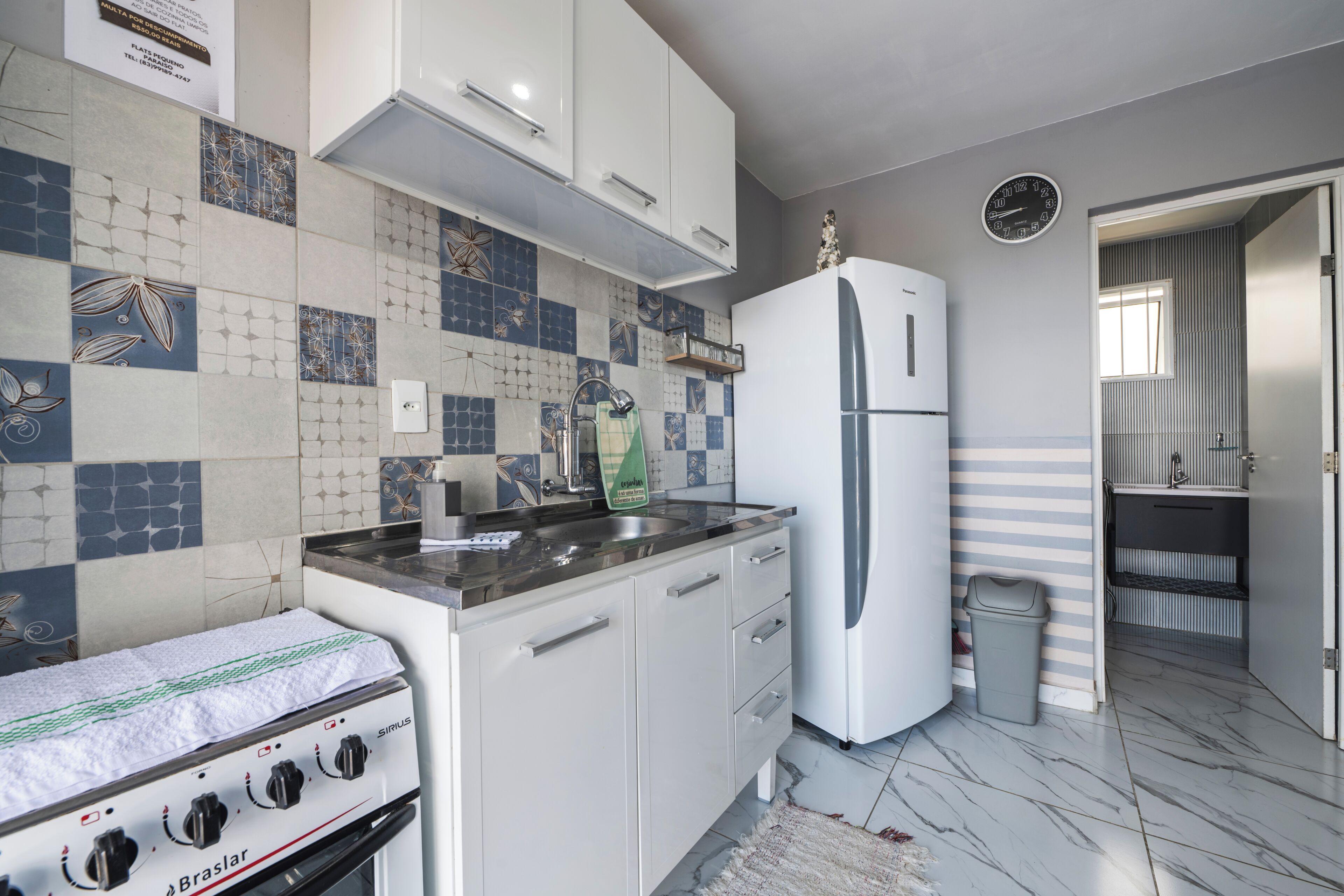 Una cocina con armarios blancos, un lavamanos de acero inoxidable, un refrigerador blanco y un salpicadero de azulejos.