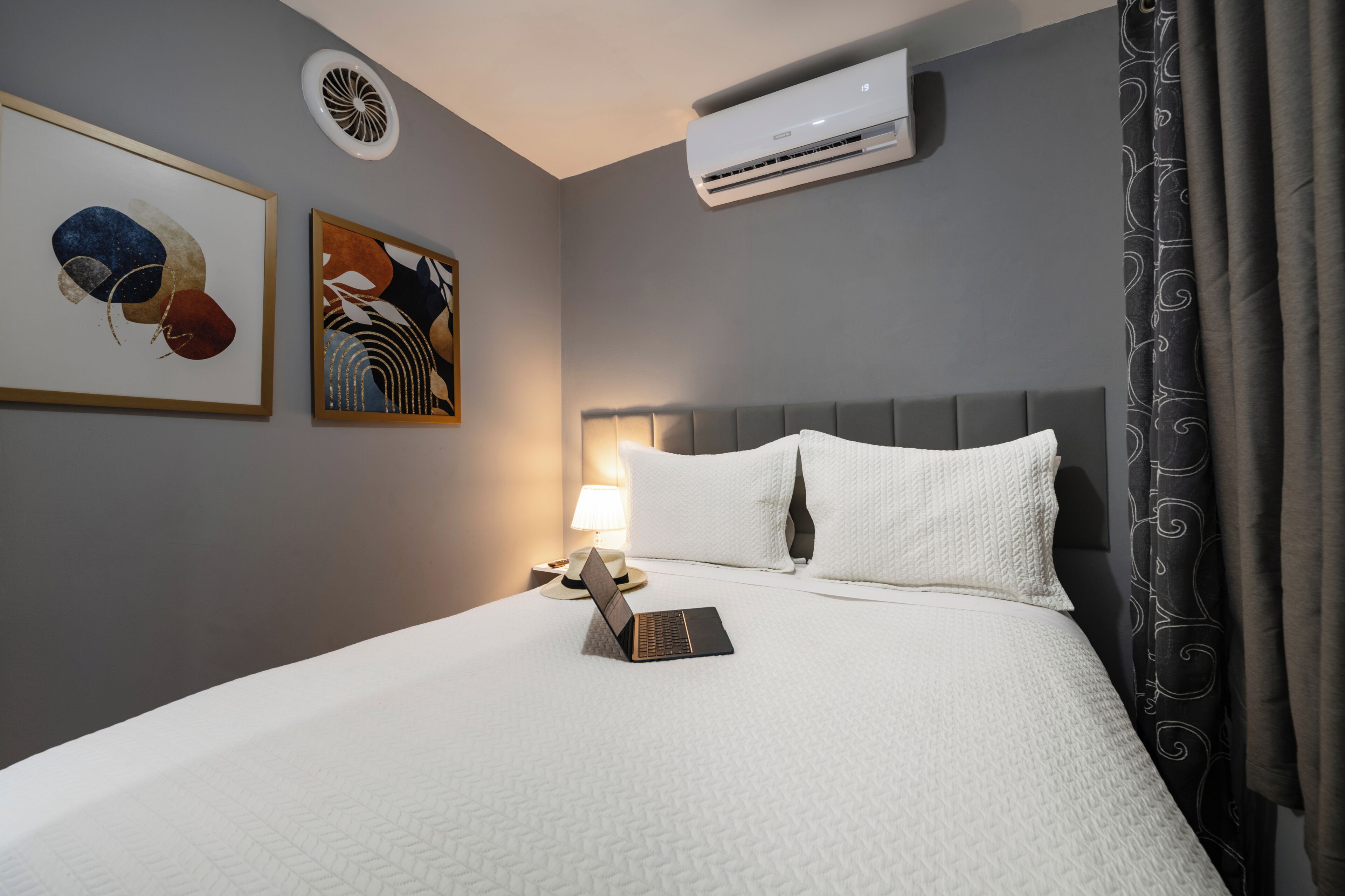 Una habitación de hotel con cama, una computadora portátil, un aire acondicionado instalado en la pared y dos obras de arte enmarcadas.