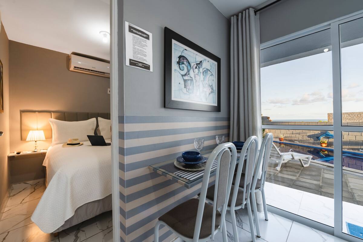 A hotel room with a bed, a desk with chairs, a view of the sea, and a framed artwork on the wall.