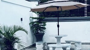 Eine Terrasse mit rundem Tisch und Bänken, umgeben von Topfpflanzen und einem großen Sonnenschirm.