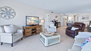 A living room with a television, white sofas, a brown leather chair, and a dining area in the background. - Gorgeous Oceanfront Condo With Double Wide Balcony & Complex Pool 🏝️🍹 (Cocoa Beach)