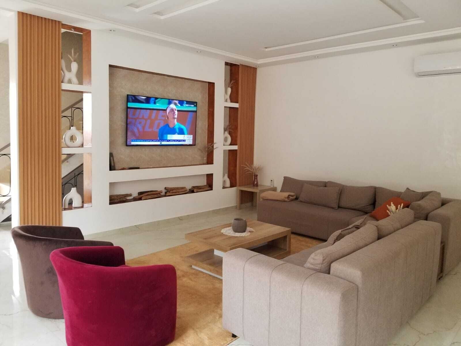 A modern living room with a large flat-screen TV, a beige sofa, a red chair, and a wooden coffee table.