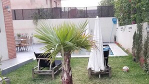 Um espaço ao ar livre pequeno, com uma palmeira, um guarda-sol branco e um banco.
