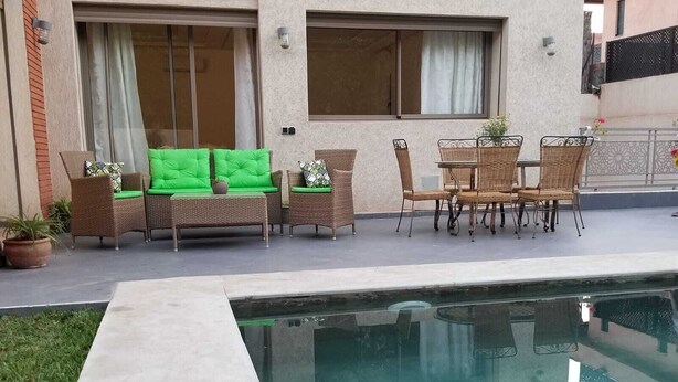 Uma área à beira da piscina com um sofá verde, cadeiras de vime e uma mesa de jantar.