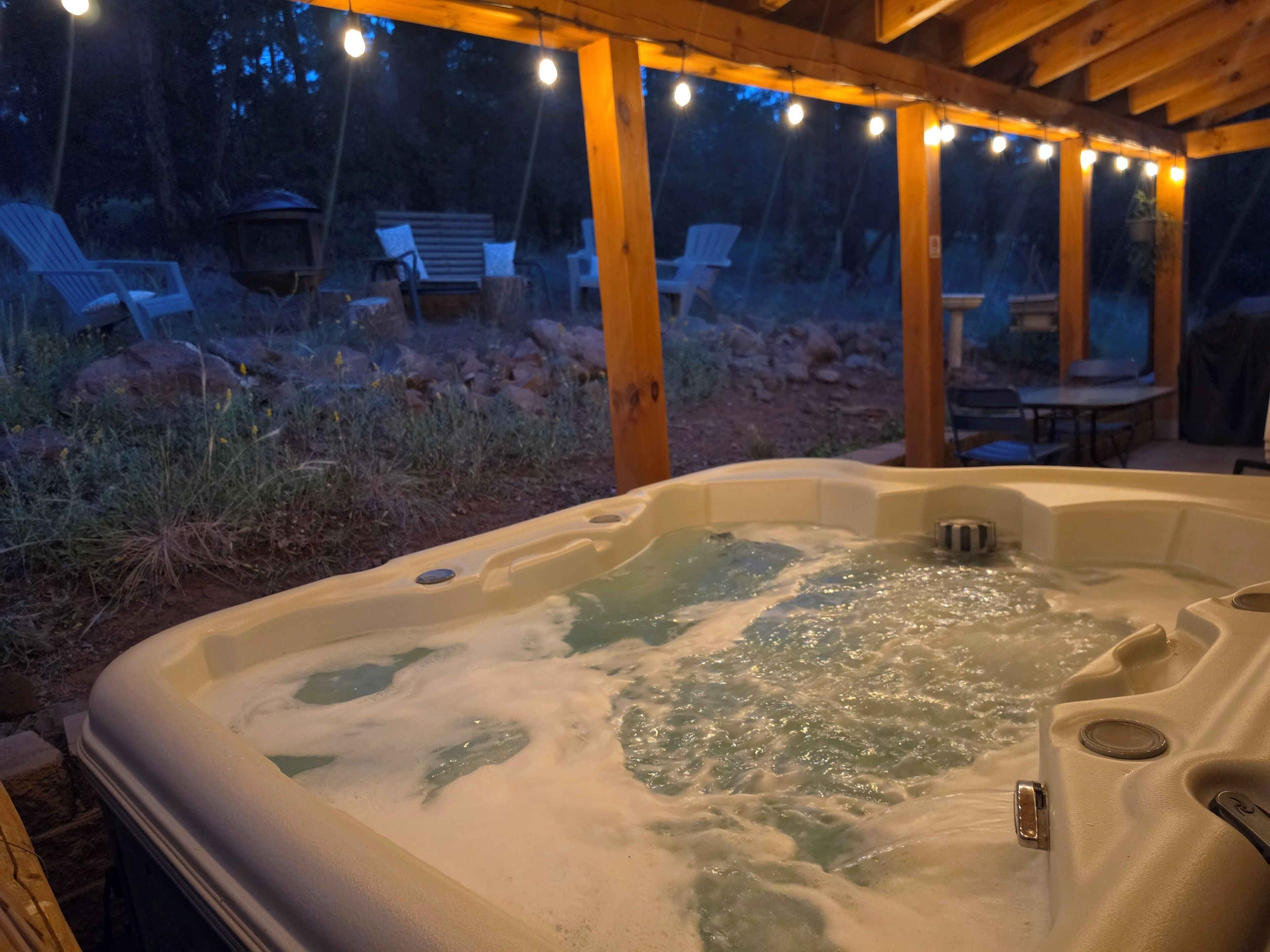 A hot tub with a wooden pergola overhead, surrounded by outdoor furniture and a natural landscape.
