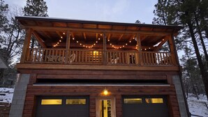 A two-story wooden cabin with a balcony, a garage, and a snow-covered ground. - Cozy Log Cabin Retreat :: Hot Tub :: Forest Views (Flagstaff)