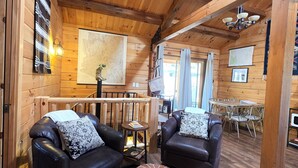 A wooden cabin interior with two leather sofas, a round ottoman, a map on the wall, and a dining area with metal chairs. - Cozy Log Cabin Retreat :: Hot Tub :: Forest Views (Flagstaff)