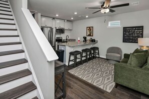 Una cocina moderna con armarios blancos, un refrigerador de acero inoxidable y un microondas. Hay una barra con taburetes y una alfombra estampada. Se ve un ventilador de techo y un cuadro enmarcado en la pared.