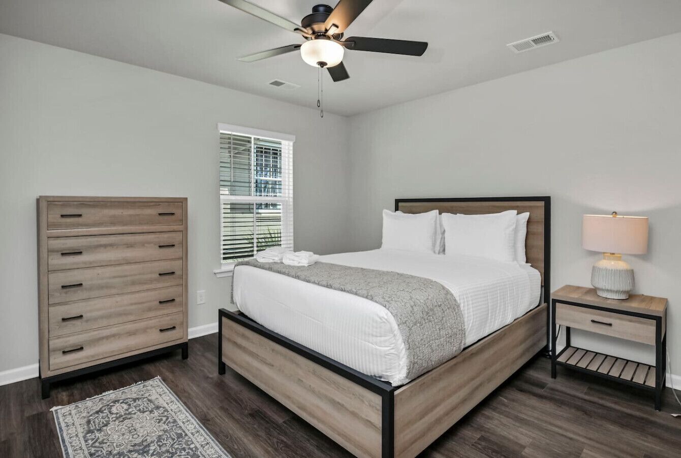 Een slaapkamer met een bed, een plafondventilator, een nachtkastje met een lamp, een dressoir en een raam met jaloezieën.