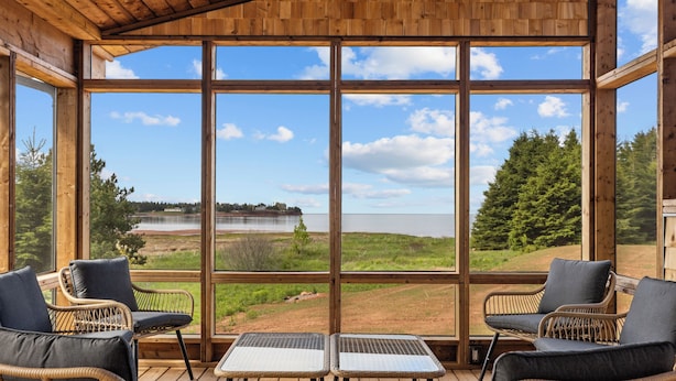 Eine Holzterrasse mit Korbstühlen und einem Tisch, große Fenster mit Blick auf einen See und Wald.