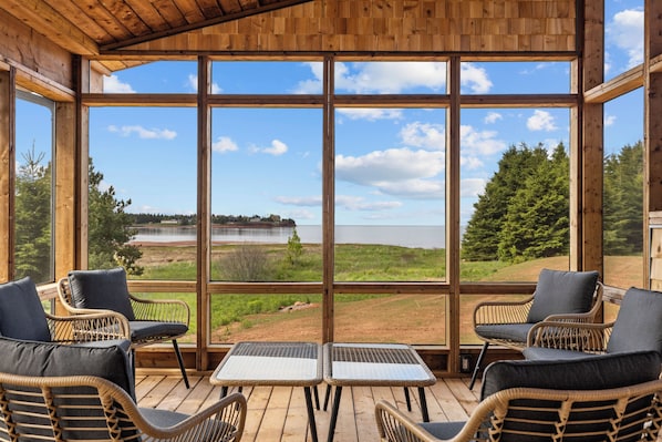 A wooden deck with wicker chairs and a table, large windows offering a view of a lake and forest.