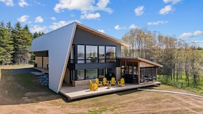 Une maison moderne à deux étages, dotée de grandes fenêtres, d’une terrasse avec des chaises jaunes et surplombant un paysage boisé.