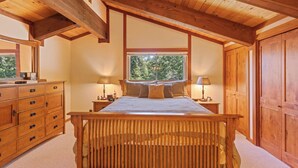 A bedroom with a wooden bed, two bedside tables with lamps, a dresser, and a large window with a view of trees. - Monthly Lease - Pine Trail at Alpine Meadows - Pet Friendly w Hot Tub at Alpine Meadows (Alpine Meadows)