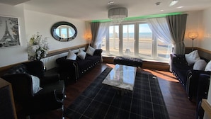 A modern living room with a large window, black sofas, a glass coffee table, and a chandelier.