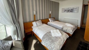 A hotel room with two beds, a window with curtains, a radiator, and a painting on the wall.