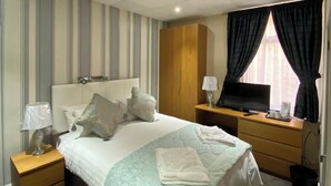 A hotel room with a bed, a wooden wardrobe, a desk with a lamp, a television, and a window with curtains.