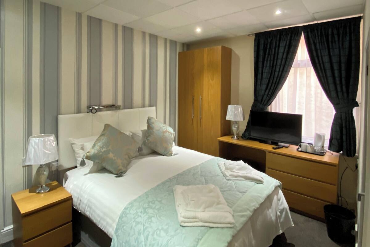 A hotel room with a bed, a wooden wardrobe, a desk with a lamp, a television, and a window with curtains.
